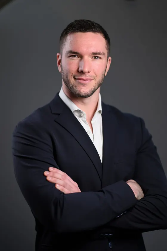 Porträt von Sascha Weihs im dunklen Sakko und weißen Hemd vor grauem Hintergrund, die Arme verschränkt.